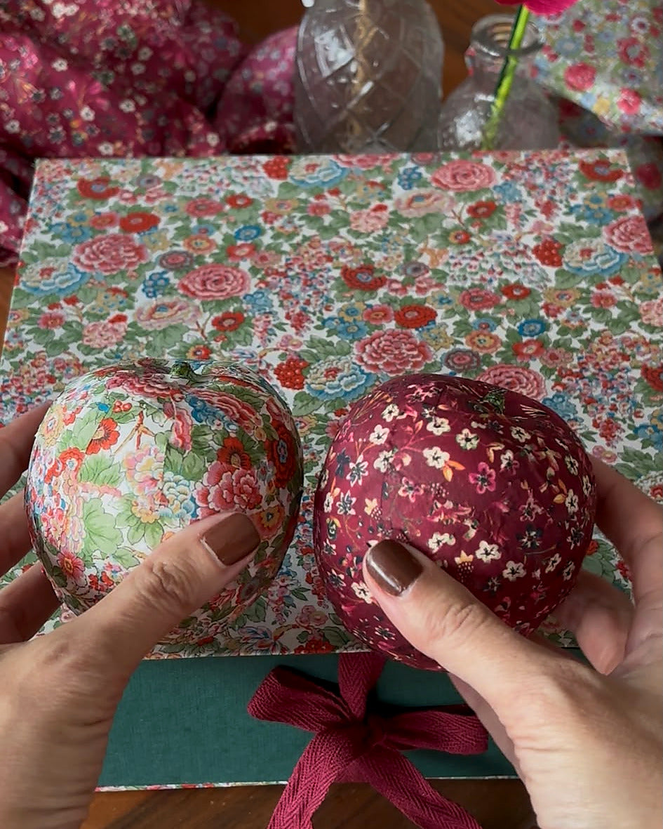 Dos manos sujetan dos manzanas decorativas terminadas con telas Liberty sobre la caja de regalo