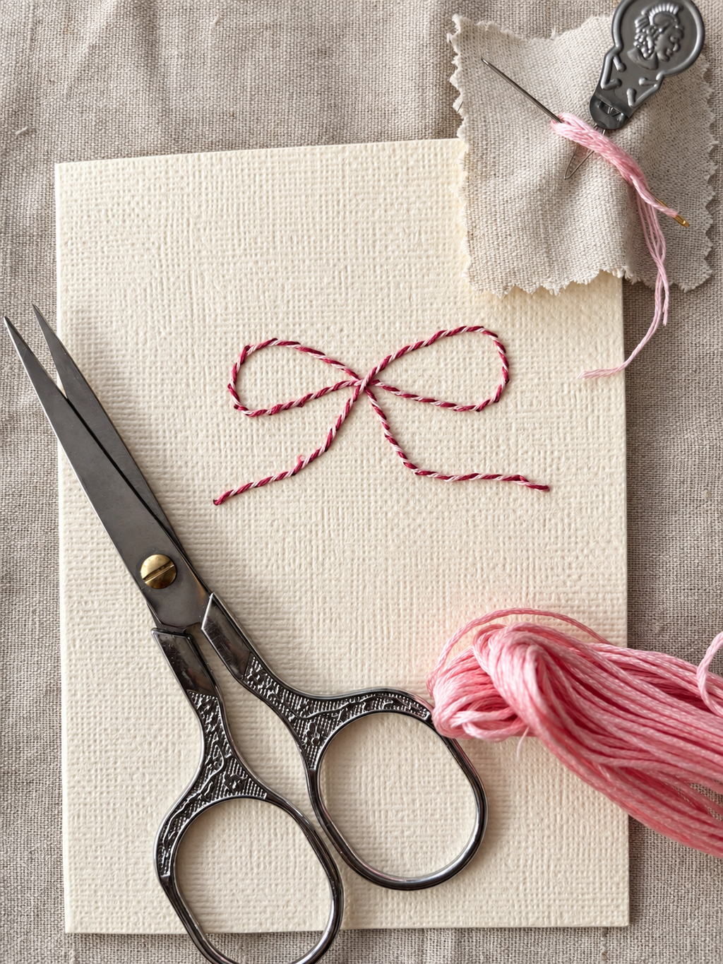 Tarjeta de papel texturizado con un lazo bordado en rosa y granate, tijeras e hilo del kit Slow Mail Atelier de Mery’s Moment.